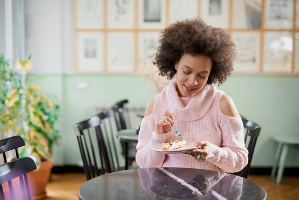 Portrait of charming positive mixed race woman in pink turtleneck sweater sitting in confectionery and eating gateau. - Photo, Image