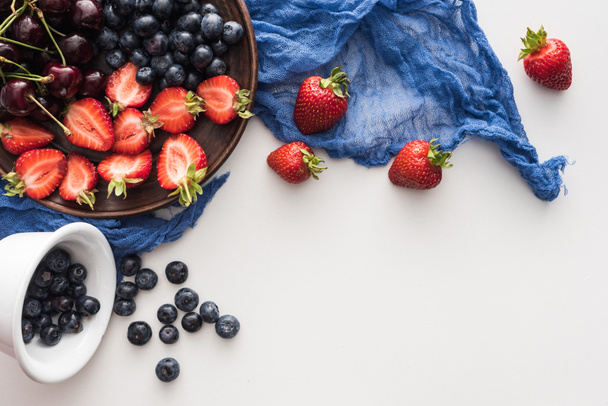 Süße Blaubeeren auf Schüssel, Kirschen und geschnittene Erdbeeren auf Teller mit blauem Tuch  - Foto, Bild