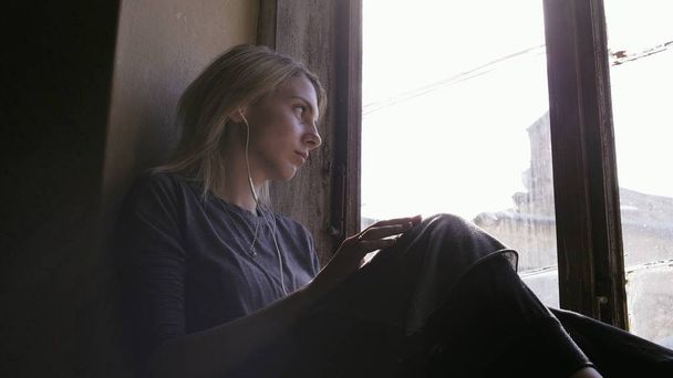 Chica se ve muy triste mientras escucha música en los auriculares por la ventana - Foto, Imagen