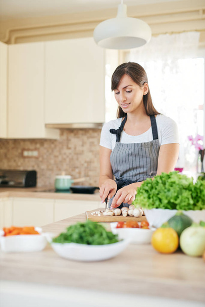 Beautiful smiling dedicated Caucasian brunette in apron standing in kitchen and chopping mushrooms. On table are lots of vegetables. Cooking at home concept. - Foto, Imagem