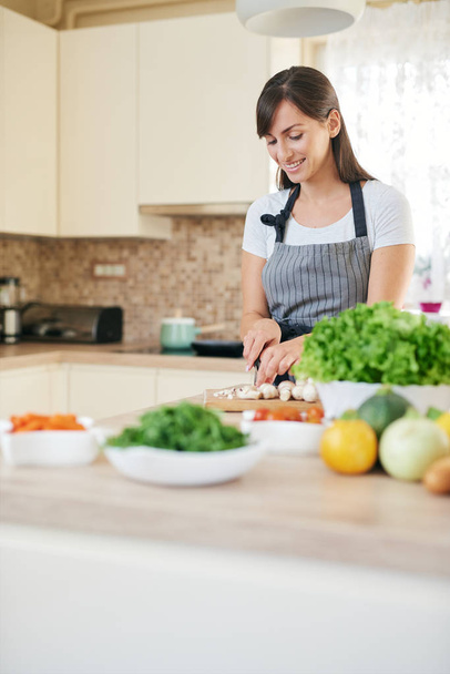 Beautiful smiling dedicated Caucasian brunette in apron standing in kitchen and chopping mushrooms. On table are lots of vegetables. Cooking at home concept. - Φωτογραφία, εικόνα