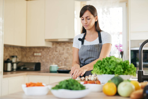 Beautiful smiling dedicated Caucasian brunette in apron standing in kitchen and chopping mushrooms. On table are lots of vegetables. Cooking at home concept. - Foto, imagen