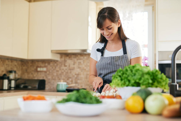 Beautiful smiling dedicated Caucasian brunette in apron standing in kitchen and chopping mushrooms. On table are lots of vegetables. Cooking at home concept. - Foto, Imagen