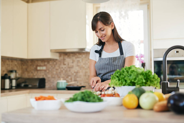 Beautiful smiling dedicated Caucasian brunette in apron standing in kitchen and chopping mushrooms. On table are lots of vegetables. Cooking at home concept. - Foto, Bild