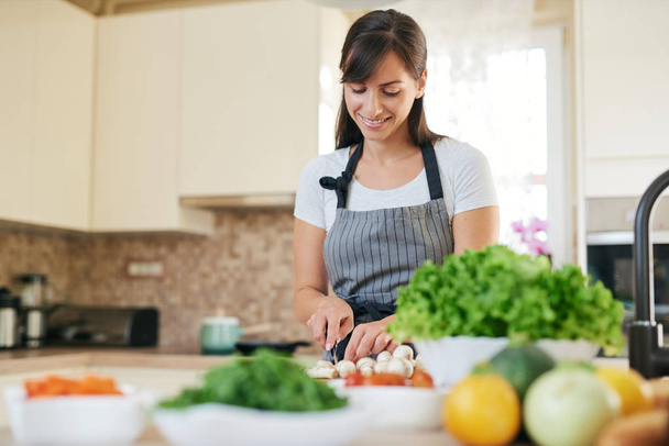 Beautiful smiling dedicated Caucasian brunette in apron standing in kitchen and chopping mushrooms. On table are lots of vegetables. Cooking at home concept. - Foto, Bild