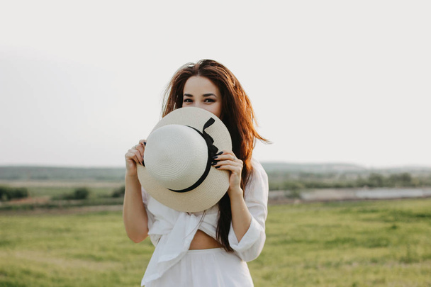 Πορτρέτο της όμορφη ανέμελη μακριά μαλλιά ασιατικό κορίτσι με λευκά ρούχα και ψάθινο καπέλο απολαμβάνει τη ζωή στο πεδίο της φύσης στο ηλιοβασίλεμα. Ευαισθησία στην έννοια της φύσης - Φωτογραφία, εικόνα