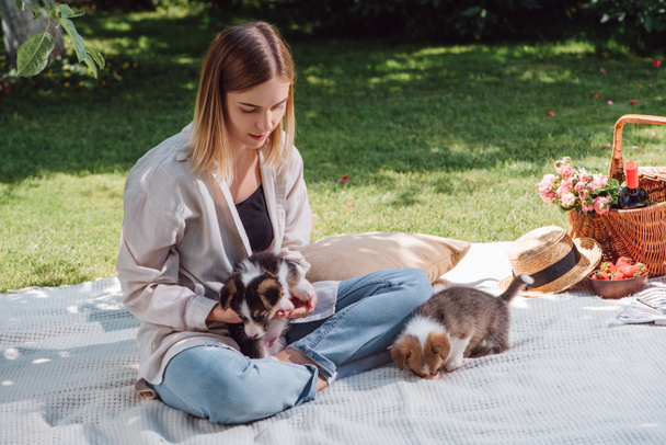 attractive blonde girl sitting on white blanket in garden and having picnic at sunny day with cute puppies - Photo, Image