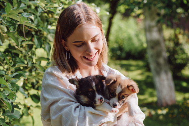 緑の木の近くの庭でかわいいウェールズコーギーの子犬を保持する幸せなブロンドの女の子  - 写真・画像