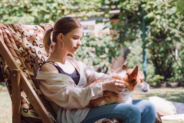blonde girl holding welsh corgi dog on knees while sitting in deck chair in garden - Photo, Image