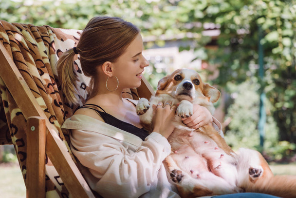 cheerful blonde girl holding welsh corgi dog while sitting in deck chair in garden - Photo, Image