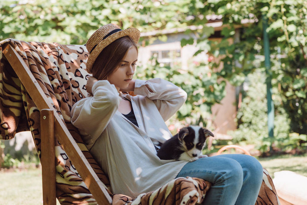 Blond dziewczyna w słomy kapelusz trzymając szczeniaka na kolana, siedząc w leżaku w ogrodzie - Zdjęcie, obraz