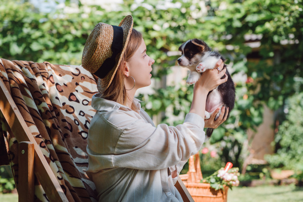 Zijaanzicht van blond meisje in stro hoed houden puppy terwijl zittend in ligstoel in de tuin - Foto, afbeelding