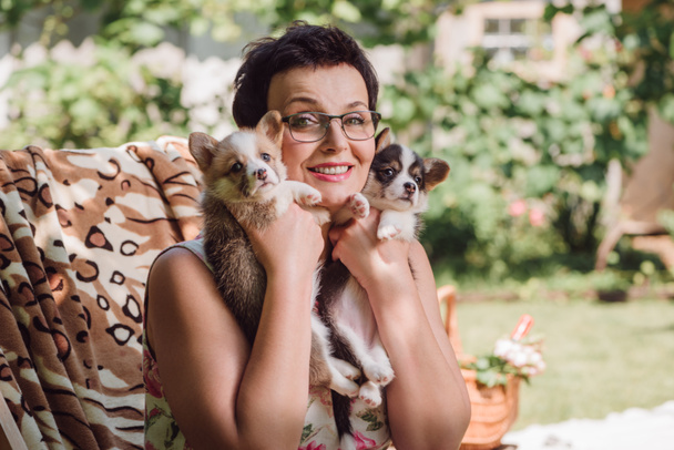 feliz morena de mediana edad mujer sosteniendo galés corgi cachorros mientras está sentado en cubierta silla en el jardín
 - Foto, Imagen