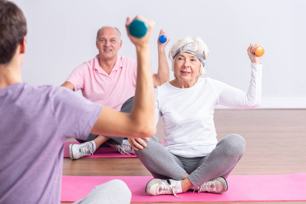 Active seniors sitting on yoga mats lifting the dumbbells during fitness classes - Foto, immagini