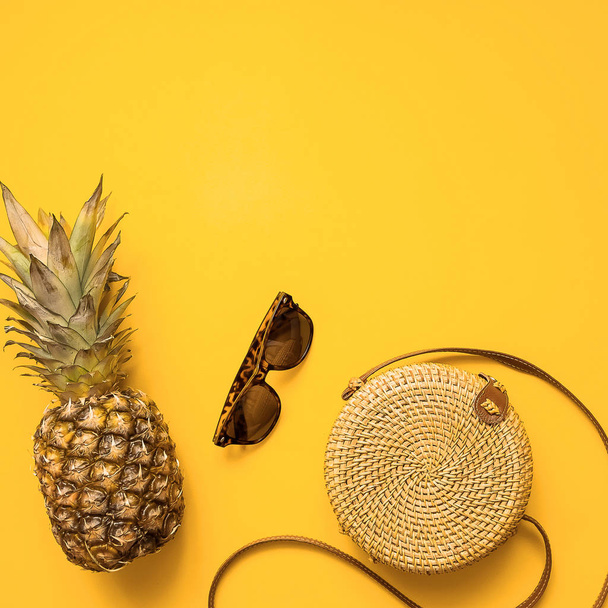 Colorful summer female fashion outfit flat lay with bamboo bag, sunglasses, pineapple over yellow background, top view. Summer fashion, holiday concept - Photo, Image