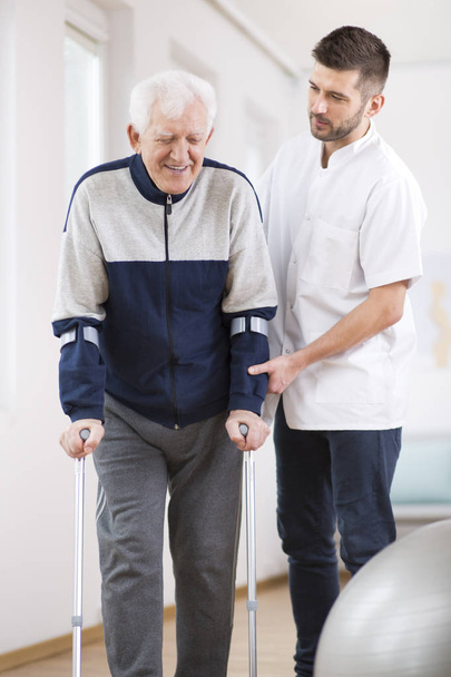 Elderly man walking on crutches and a helpful male nurse supporting him - 写真・画像