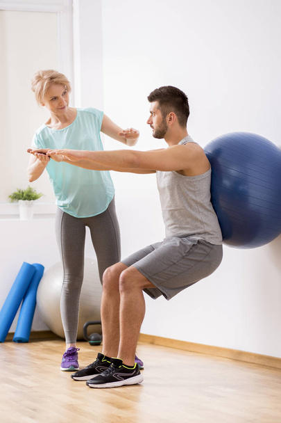 Young man with back injury exercising with blue gymnastic ball during appointment with female physiotherapist - Foto, Imagen