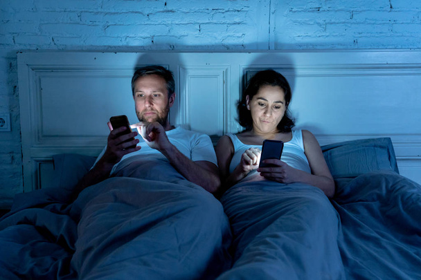 Young couple in bed late at night using smart phones obsessed with games, social media and apps ignoring each other in relationship communication problems and internet mobile addiction concept. - Photo, image