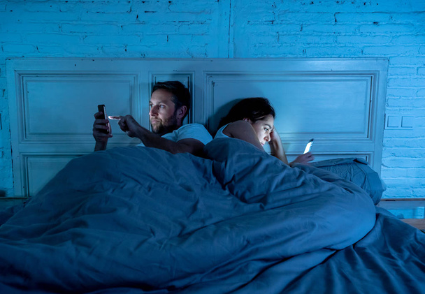 Couple affected by technology addiction ignoring at each other in apathy and anger lying in bed using their smart phones late at night in darkness in relationship issues and internet mobile addiction. - Φωτογραφία, εικόνα