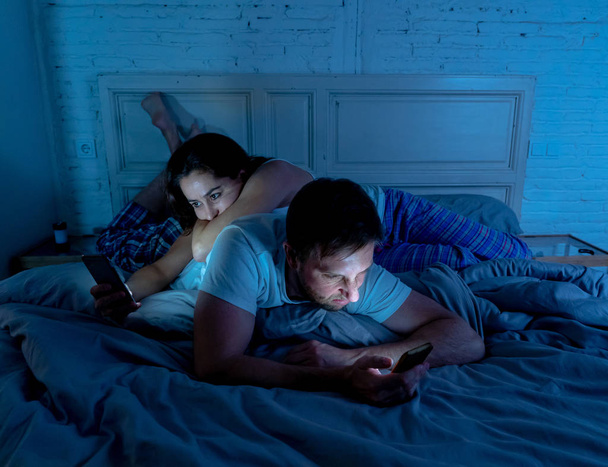 Sad man and woman married couple using their smart mobile phone in bed at night ignoring each other as strangers in relationship and communication problems and internet social network addiction. - Photo, image