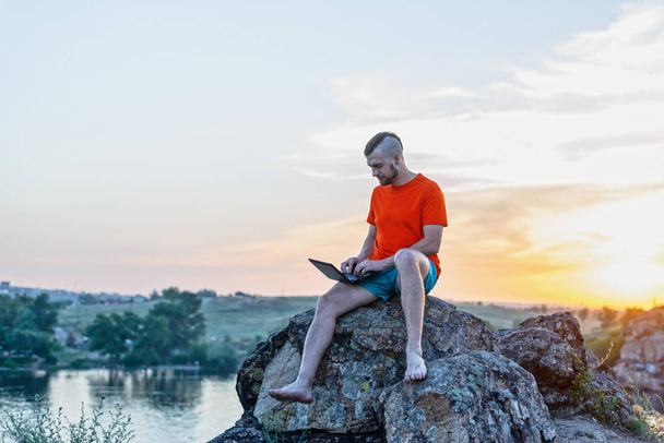 A man working on laptop sitting on the peak of a mountain at sunset. Technology, internet, staying online. - Zdjęcie, obraz