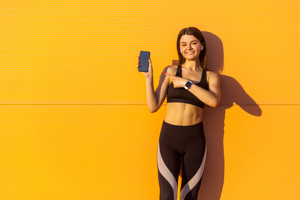 Young satisfied beautiful sporty woman in black sportswear pointing finger at smartphone in hand while standing near orange wall background in sunshine  - Foto, Bild