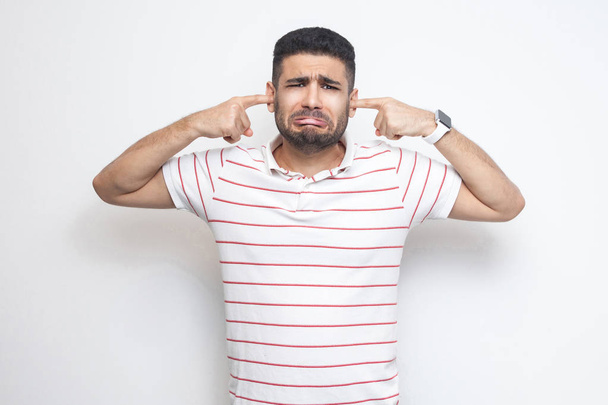 sad confused bearded young man in striped t-shirt looking at camera and putting his fingers on ears isolated on white background - Foto, afbeelding
