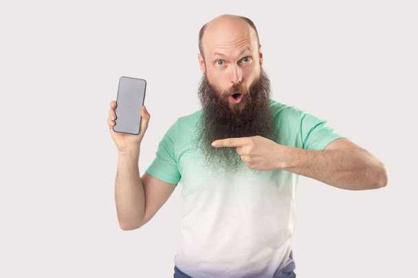 shocked middle aged bald bearded man pointing at smartphone empty screen and looking at camera with surprised face on grey background. - Foto, immagini
