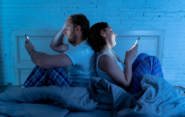Couple in bed using phones, husband frustrated upset and unsatisfied while internet addict wife ignores him and enjoys mobile phone in social network addiction and relationship domestic problems. - Photo, image