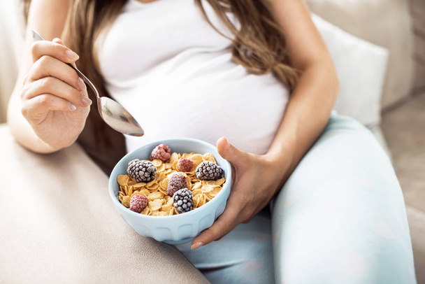 Закрытие счастливой беременной женщины, которая ест цералы с фруктами на завтрак в постели дома
 - Фото, изображение