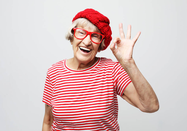 Lifestyle, emotion and people concept: Funny old lady wearing red hat and eyeglasses showing ok sign - Foto, Bild