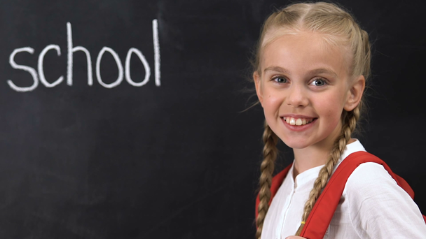 Little girl with rucksack smiling at camera, school word written on blackboard - Footage, Video