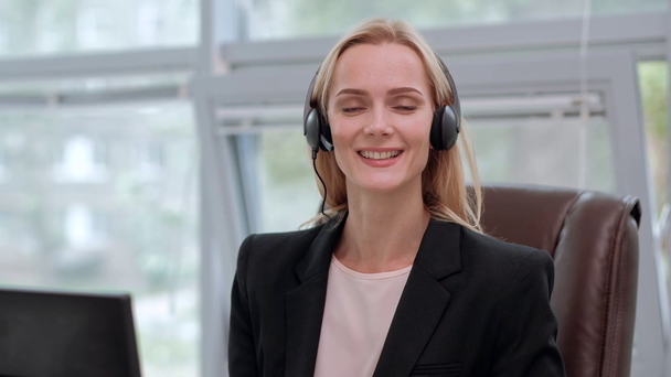 A young attractive woman wearing a black jacket in a black jacket is sitting at her desk in a executive chair. Holds a video conference in headphones with a microphone. - Filmati, video