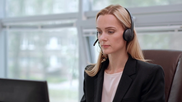 Eine junge attraktive Frau in schwarzer Jacke sitzt in einem Chefsessel an ihrem Schreibtisch. hält eine Videokonferenz im Kopfhörer mit Mikrofon ab. - Filmmaterial, Video