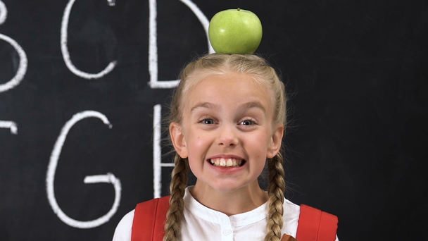 Funny little girl standing with apple on head, alphabet written on blackboard - Video
