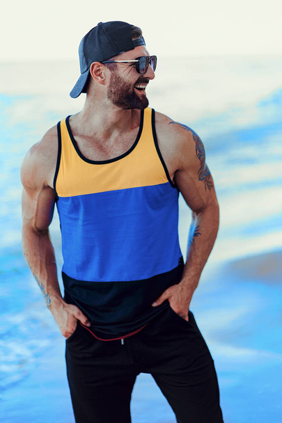 Bearded tattooed man at the resort in a sportive shirt, cap and sunglasses on the sea background - Foto, Imagen
