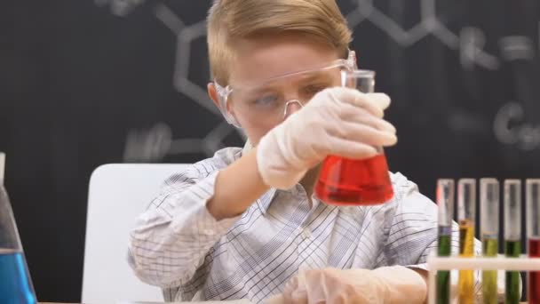 Schoolboy looking at flask with red liquid, writing reaction data in copy-book - Filmati, video