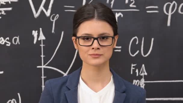 Female scientist taking off eyeglasses looking at camera, formulas on blackboard - Video