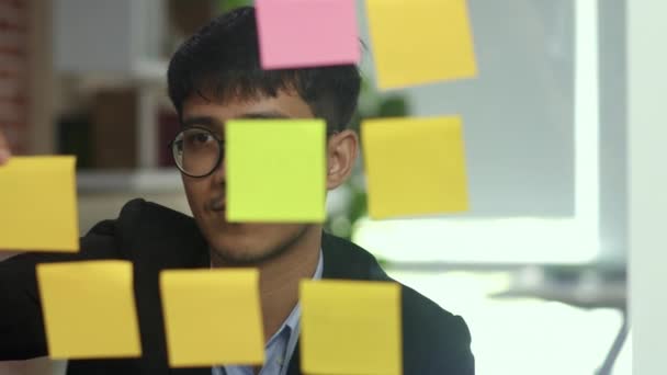 Asian creative man stick a sticky note on glass board. Young professional business male write information, strategy, reminder on paper, business situation, startup in Loft office concept. Slow motion. - Footage, Video