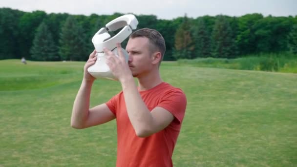 Egy férfi hozza a VR headset a fejét egy parkban, és körülnéz. Egy fiatal srác egy piros póló és egy sisak tapasztalható virtuális valóság a háttérben a természet. Dinamikus lövöldözés. 4k felvételek. - Felvétel, videó