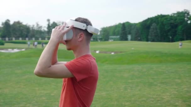 Ein Mann setzt sich in einem Park ein Headset auf den Kopf und schaut sich um. Ein junger Mann in rotem T-Shirt und Helm erlebt Virtuelle Realität vor dem Hintergrund der Natur. Dynamisches Schießen. 4k Filmmaterial. - Filmmaterial, Video