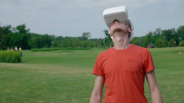 Een man met behulp van een vr-headset in het Park en rond te kijken. Een jonge kerel in een rood T-shirt en een helm ervaart Virtual Reality tegen de achtergrond van de natuur. Een mens aan de rechterkant in het frame. 4k-beeldmateriaal. - Video
