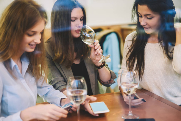 Attractive caucasian female bloggers drinking white wine in cafeteria for take little rest while posting new publication on profile via application on cellular phone, millennial women using cellphones - Photo, Image