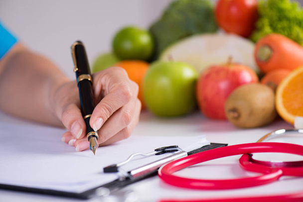 Υγιεινός τρόπος ζωής, φαγητό και διατροφική έννοια. Κοντινό επάνω γιατρός γυναίκα χέρι κρατώντας στυλό σε λίστα ελέγχου με φρέσκα λαχανικά και φρούτα με στηθοσκόπιο που βρίσκεται σε επιφάνεια εργασίας στο παρασκήνιο. - Φωτογραφία, εικόνα