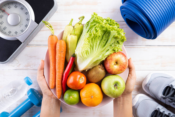 Healthy lifestyle, food and sport concept. Top view of woman hand holding plate of fresh vegetables and fruits with athlete's equipment Weight Scale blue dumbbell, sport water bottles on white wooden background. - Фото, изображение