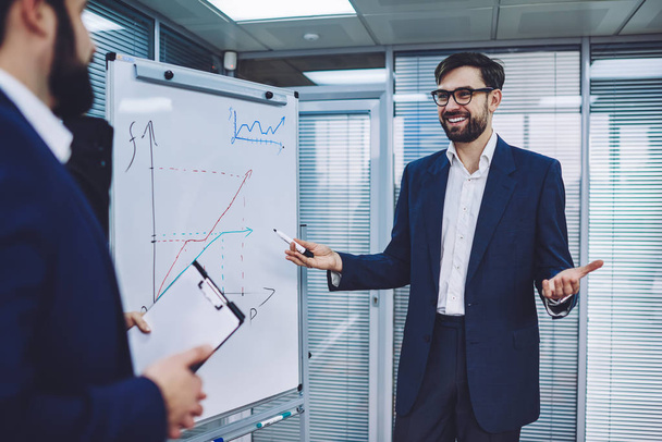 Successful male entrepreneur in optical spectacles for vision correction standing near flip chart with economic graphics and communicating with business partner about main topic of develope company - Foto, Bild