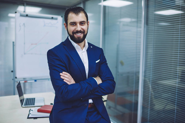 Portrait of successful male boss dressed in elegant blue suit standing at own office room with crossed arms and smiling at camera during working time, happy man enjoying business lifestyle indoors - Fotoğraf, Görsel