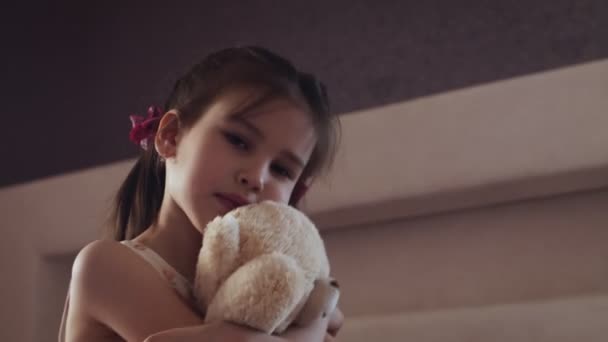 Little girl flowered dress sits on the bed and hugs teddy bear. - Video
