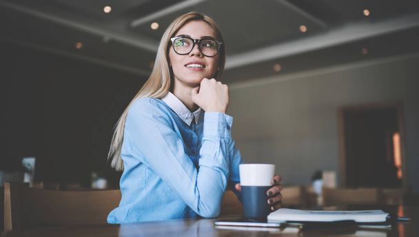 Attractive hipster girl thoughtful looking away while sitting at university table with cup and thinking about course work, pensive female student in stylish spectacles pondering on college homework - Foto, Imagen