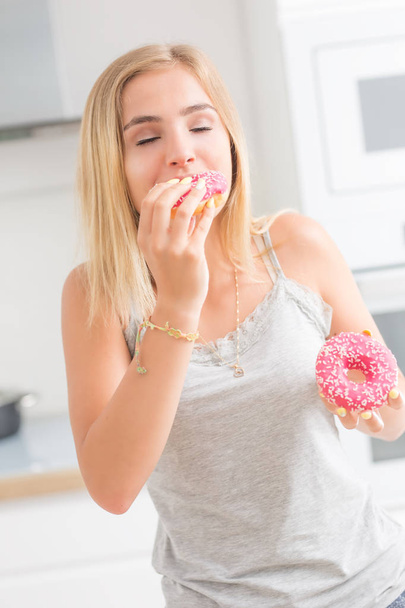 Young blonde girl eats pink donuts in home kitchen with taste em - Fotografie, Obrázek